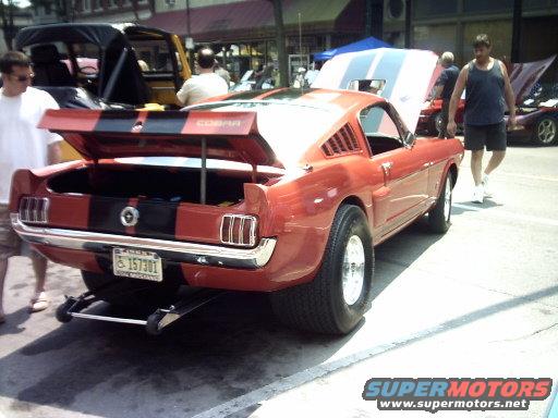 Red 1965 Ford Mustang fastback with black racing stripes, trunk open, license plate 157301