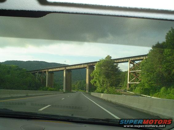 view from car driving on highway towards a tall railroad bridge crossing over the road, surrounded by trees and hills