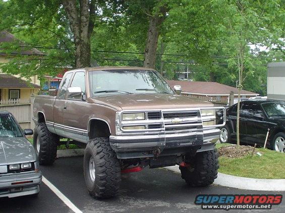 Lifted brown Chevrolet pickup truck with large tires parked next to a silver car and a black car