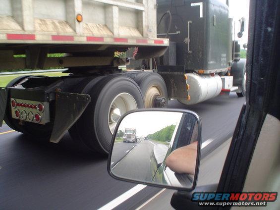 View from car window shows side of semi-truck trailer and reflection of another semi-truck in side mirror