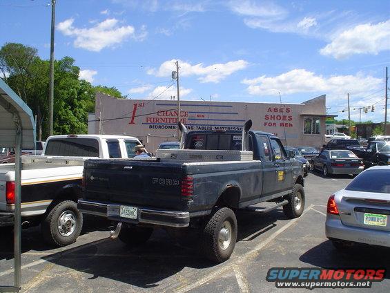 Dark Ford pickup truck with dual exhaust stacks and a toolbox in the bed, parked next to a white pickup truck