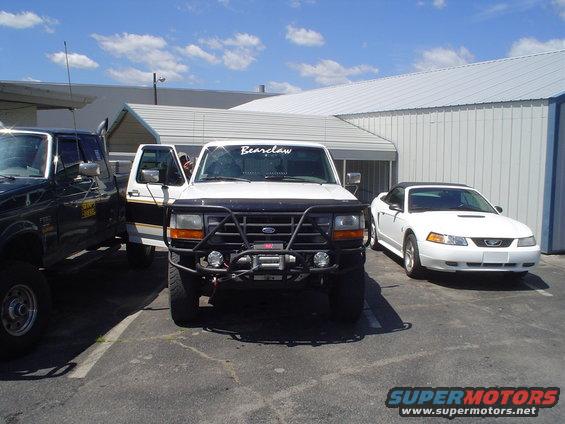 White Ford truck with bull bar and winch, parked next to a black truck and a white Ford Mustang convertible