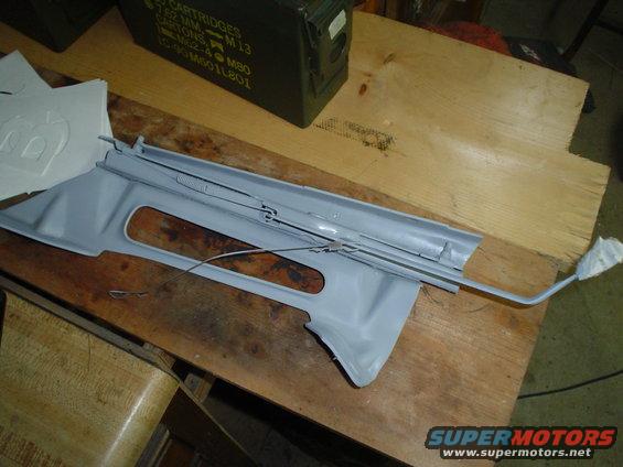 Gray plastic car interior trim piece with metal cables attached, resting on a wooden surface
