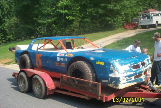 Blue stock car with orange roll cage on a trailer, people standing nearby