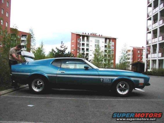 Blue 1971 Ford Mustang Mach 1 parked, man loading trunk. Buildings in background