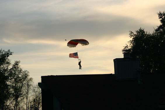 Skydiver descends with American flag against a cloudy sky, over a dark roof and trees