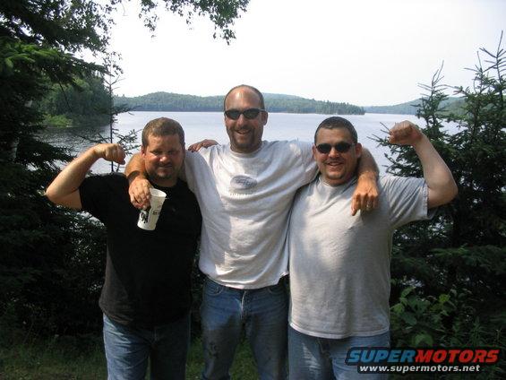 Three guys posing by a lake, flexing their biceps