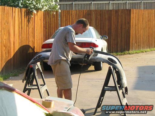 Man sanding a white car's rear bumper on sawhorses