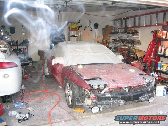 Red sports car with hood covered in plastic, parked next to a white car in a garage. Bodywork is in progress