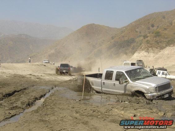 Silver pickup truck stuck in mud with other vehicles in the background on a dusty hill