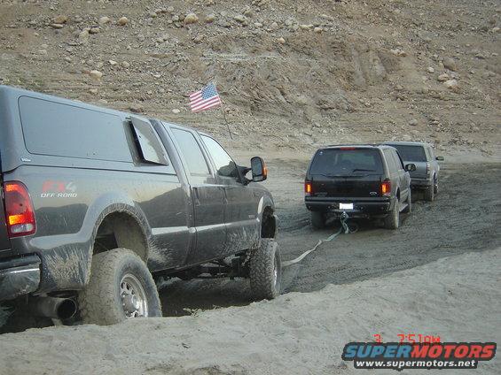 Dark pickup truck with a camper shell is towing two SUVs through a sandy, off-road area. An American flag is attached to a pole on the side of the pickup