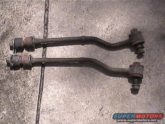 Two dark metal suspension control arms with rubber bushings and bolts, resting on a concrete surface