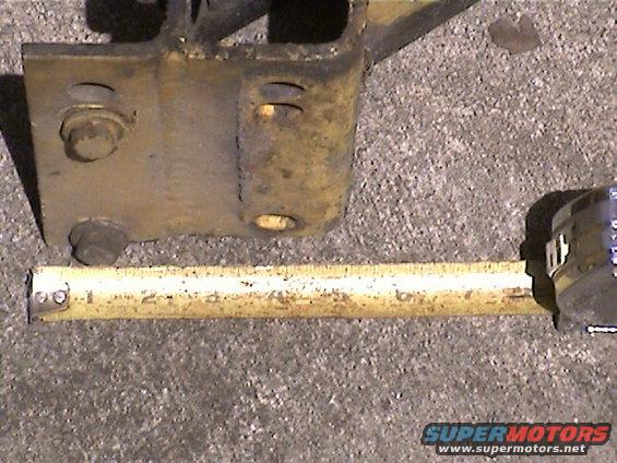 Rusty yellow metal bracket with bolts next to a yellow measuring tape on a concrete surface