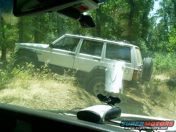 White Jeep Cherokee on a dirt trail, angled uphill. Spare tire mounted on the rear