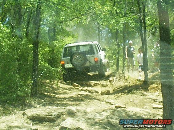 SUV driving up a rocky, wooded trail with people watching