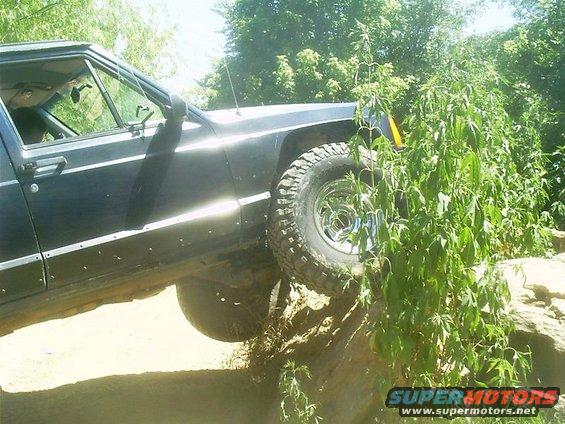 Black Jeep Cherokee XJ with off-road tires climbing over rocks and brush