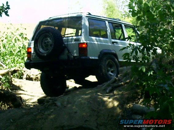White Jeep Cherokee XJ with spare tire on rear, driving on a dirt trail