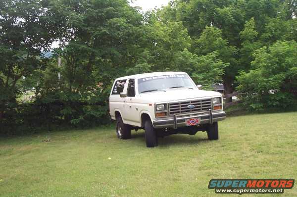 White Ford Bronco parked on grass with trees in background. Has a Confederate flag plate on the front