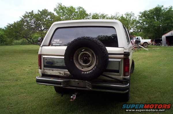 1985 Ford Bronco rear with spare tire mounted externally. Another white pickup truck is visible in the background