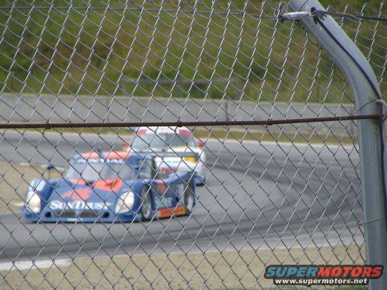 Blue and orange race car with SunTrust livery on a track, seen through a chain-link fence