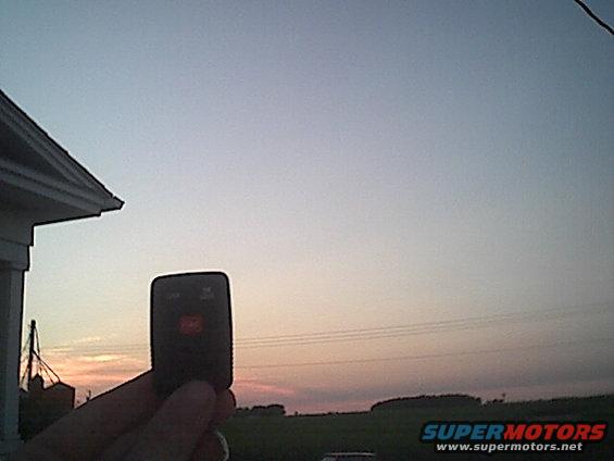 Hand holding a car key fob with two buttons, silhouetted against a sunset sky with power lines and a distant treeline