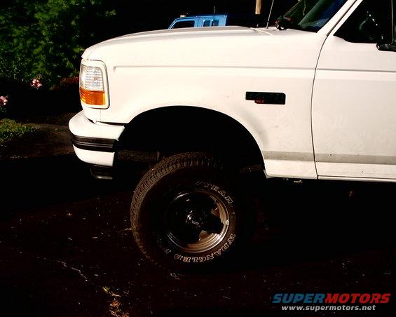 1996 Ford Bronco front fender, wheel, and tire. Blue vehicle in background
