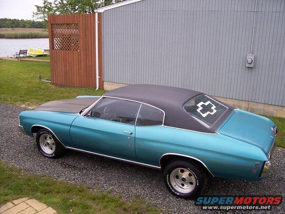 Teal coupe with black vinyl roof parked by a building near water. Has a white cross decal on rear window