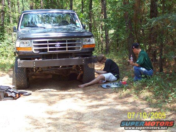 Dark gray Ford truck with aftermarket bumper, front view, with two men working on it in a wooded area