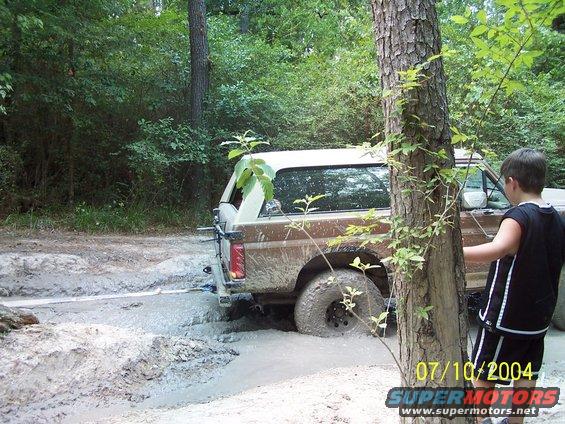 Brown 1987 Ford Bronco stuck in mud, rear view, with a boy standing nearby