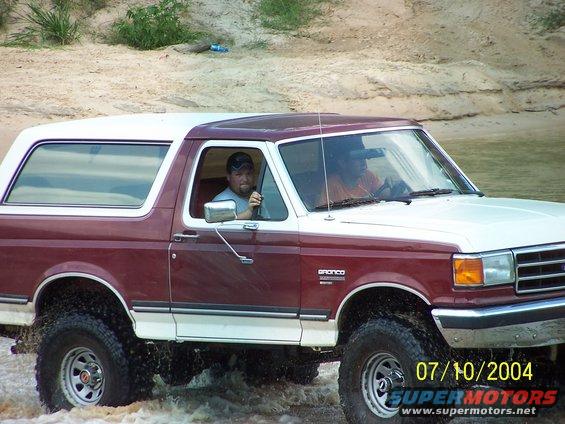 Maroon and white 1987 Ford Bronco driving through water, driver visible