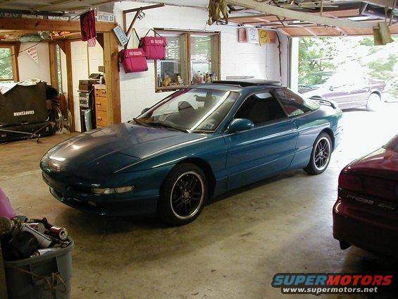 Teal coupe with a sunroof parked in a garage next to a red car. Another car is visible in the background