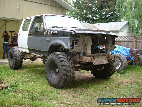 Modified black and white Ford truck with oversized tires, missing front end, parked on grass