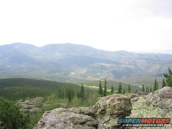 Mountain landscape with pine trees and rocks in the foreground. A watermark reads SUPERMOTORS