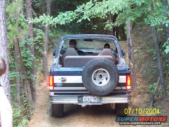 Ford Bronco driving on a dirt trail with trees on both sides. Two people are inside