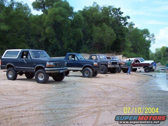 Line of lifted Ford Broncos and trucks parked on a sandy beach with trees in the background. 1987 Ford Bronco is visible