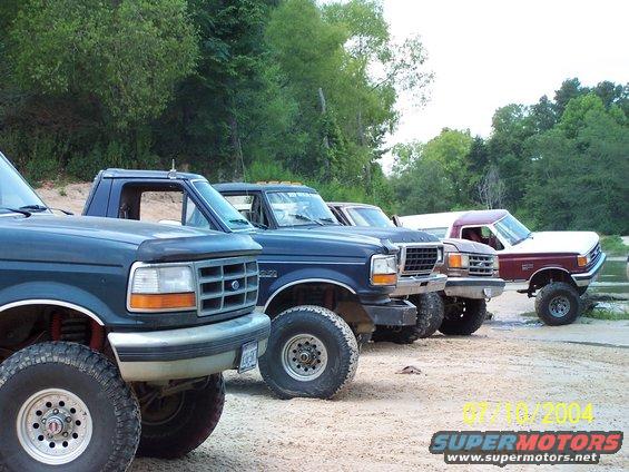 Four lifted Ford trucks parked in a line, front-left to rear-right. The closest truck is dark blue with a gray bumper. The second truck is dark blue, the third is brown, and the fourth is white with a maroon cab