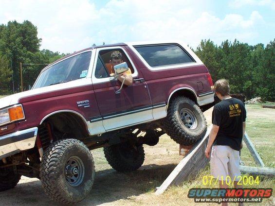 Maroon and white 1987 Ford Bronco with lifted suspension driving over an obstacle