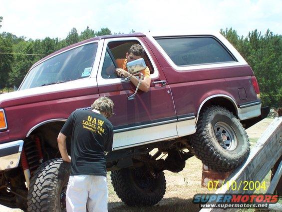 1987 Ford Bronco with two people, one driving, one looking at the front wheel while on a wooden obstacle