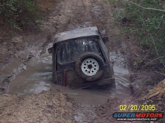 Jeep-like vehicle stuck deep in a muddy ditch, spare tire visible on the back