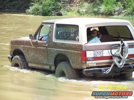 Brown 1987 Ford Bronco driving through muddy water, rear view