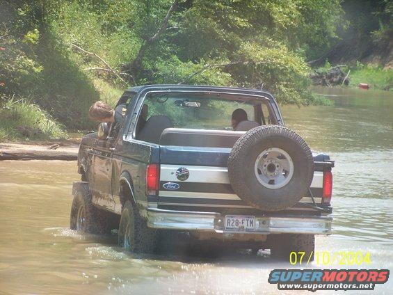 1987 Ford Bronco driving through shallow water, spare tire mounted on rear door. License plate R28-FTM