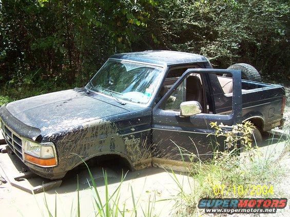 Mud-covered black Ford Bronco stuck in mud, driver's door open, spare tire visible in the bed