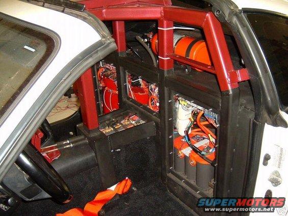 Interior of a vehicle with a red roll cage, custom panels holding batteries and wiring, and an orange tank