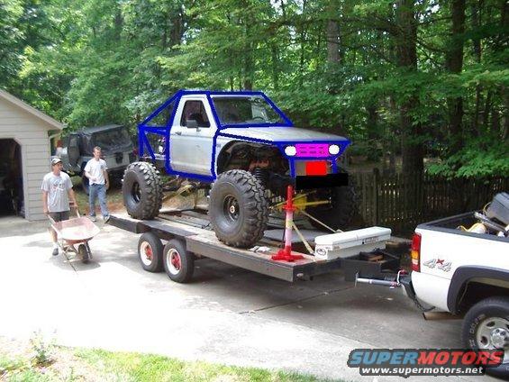 Silver pickup truck with blue roll cage on trailer, large tires, purple headlights, red grille