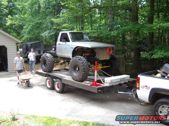 Silver pickup truck chassis with large tires on a trailer, being loaded by two men. Another SUV is parked nearby