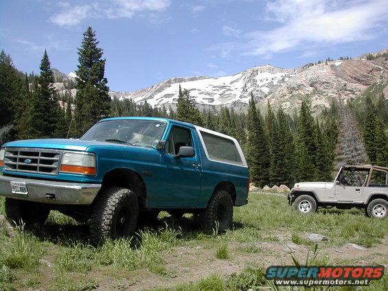 Teal 1994 Ford Bronco parked in a grassy field with a Jeep Wrangler behind it. Snowy mountains and pine trees in the background