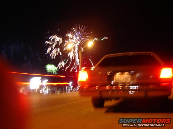 Rear view of a tan sedan with taillights illuminated, driving at night with fireworks in the background