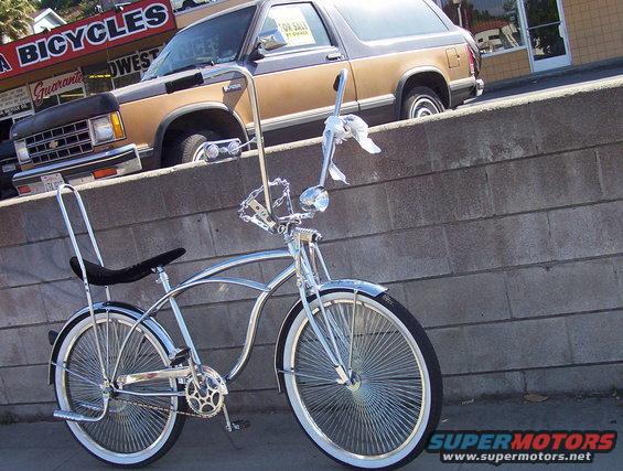 Chrome lowrider bicycle parked in front of a brick wall, with a two-tone pickup truck behind it