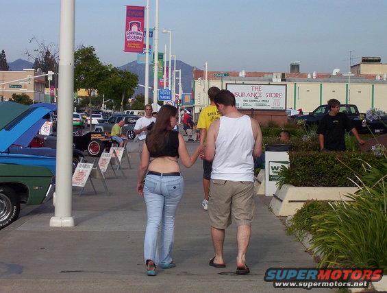 People walk past parked cars at an outdoor event, with buildings and mountains in the background