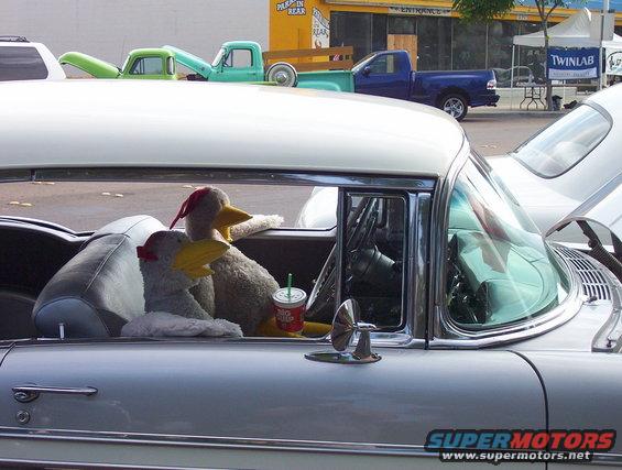 Two stuffed ducks with red bows sit in the front seats of a classic car, with a drink cup between them. Other vehicles are visible in the background